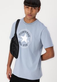 Tricou din bumbac de culoare albastru deschis, cu un grafic circular alb ce conține o stea și textul "Converse All Star". Geantă neagră crossbody pe umăr.