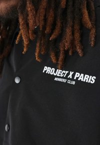 Camisa negra de botones con un logo blanco que dice "PROJECT X PARIS MEMBERS' CLUB". El tejido parece suave con un acabado mate.