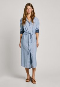 Abito camicia a righe blu e bianche, con bottoni, cinte in vita con una cintura. Presenta maniche lunghe arrotolate e una lunghezza media.