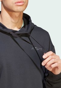 Sudadera negra con logo bordado, cordón ajustable y una textura de tela suave. Presenta un corte holgado y un bolsillo canguro frontal.