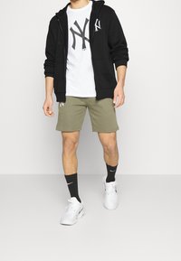 Felpa nera con zip, t-shirt bianca con grafica grande, pantaloni corti verde oliva, calze nere con logo bianco e sneakers bianche.