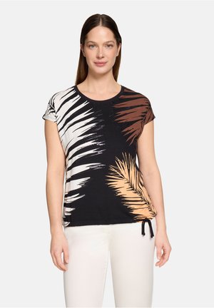 Kvinde iført en sort top med korte ærmer med hvide, brune og beige bladmønstre, kombineret med hvide bukser, stående mod en ensfarvet baggrund.