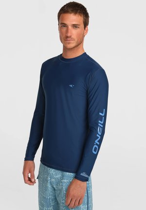 Man met een marineblauw, lang mouw O'Neill rashguard en lichtblauwe zwemshorts met patroon tegen een effen lichte achtergrond.