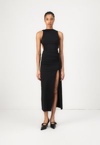 Melnā formā pieguļoša kleita ar augstu šķēlumu un krokojuma detalizāciju, ar bez piedurknēm un atvērtu muguru. Uzsvērta ar melniem papēžiem.