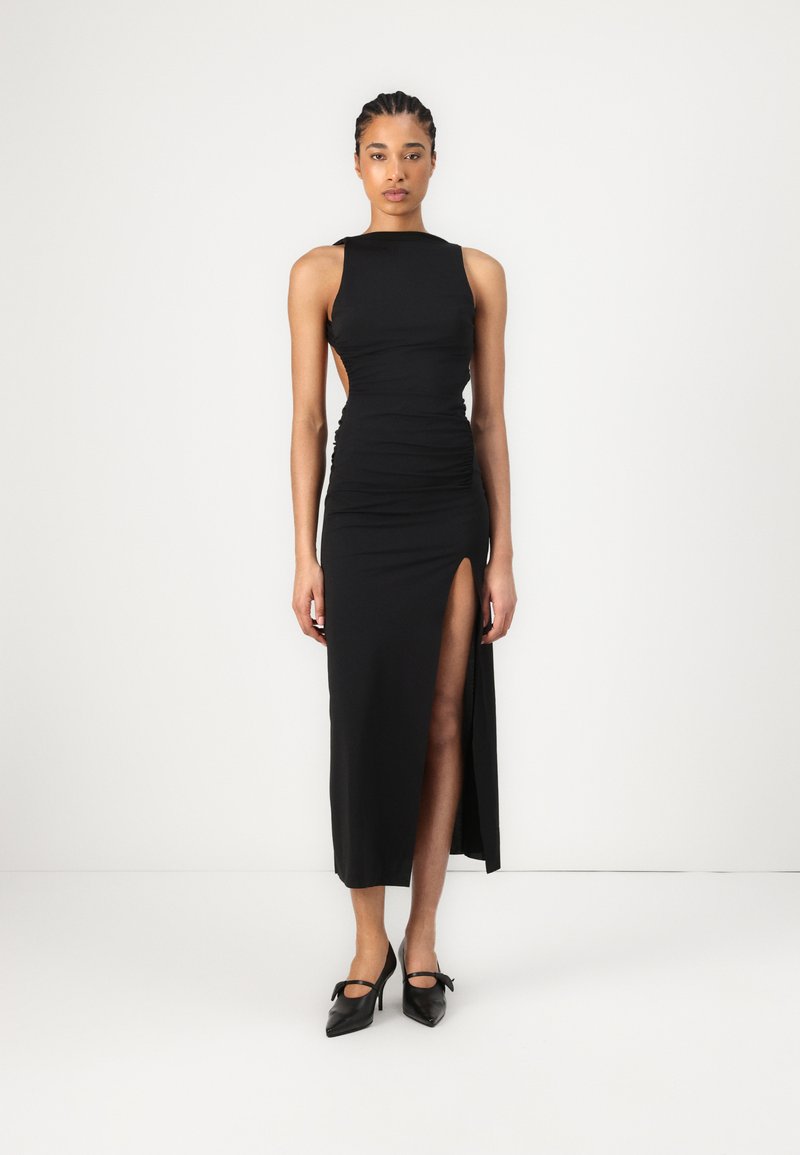 Melnā formā pieguļoša kleita ar augstu šķēlumu un krokojuma detalizāciju, ar bez piedurknēm un atvērtu muguru. Uzsvērta ar melniem papēžiem.