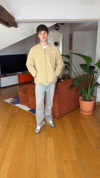 Noormees, kes kannab beeži jopet ja helesiniseid pükse, seisab elutoas, kus on oranž diivan, televiisor, puidust põrand ja suur potitaim.