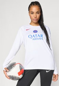 Maillot à manches longues blanc avec le logo bleu "QATAR AIRWAYS" et le swoosh rouge de Nike. Leggings noirs de Nike. Le joueur tient un ballon de football à motifs.