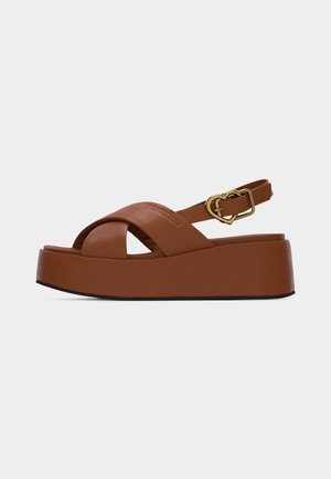 Brown platform sandal with crisscross straps and a gold heart-shaped buckle on the ankle strap, flat black sole edge.