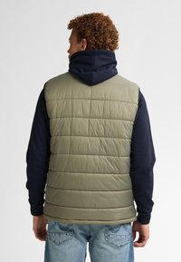 Grüne Pufferweste mit horizontalen Nähten, getragen über einem marineblauen Hoodie. Legere Jeans ergänzen den geschichteten Look.
