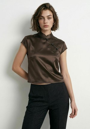 Femme aux cheveux courts bruns portant un haut chinois brillant marron avec des fermetures à boutons nœuds et un pantalon noir taille haute, debout contre un mur blanc.