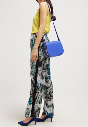 Kvinde iført gul ærmeløs top, mønstrede bukser med vide ben, blå hæle og en blå crossbodytaske, der står sidelæns.