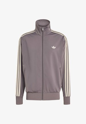 Grå träningsjacka med hög krage, hel dragkedja och elastiska ärmmuddar. Har vita ränder på ärmarna och en liten logotyp på bröstet.