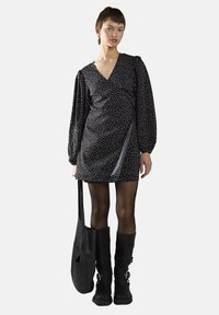 Musta pilkullinenWrap-mekko, jossa on pitkät hihat ja V-pääntie, yhdistettynä mustiin polvipitkiin saappaisiin ja mustaan olkalaukkuun.