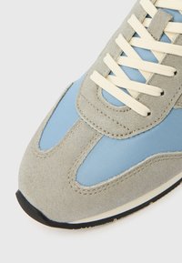 Baskets en daim gris avec des panneaux en tissu bleu clair, des lacets crème et une semelle en caoutchouc noire. Présente des accents cousus sur la tige.