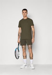 Mann med tatoveringer iført olivengrønn treningstøy og hvite sneakers, som holder en blå og svart tenniskølle mot en ensfarget bakgrunn.