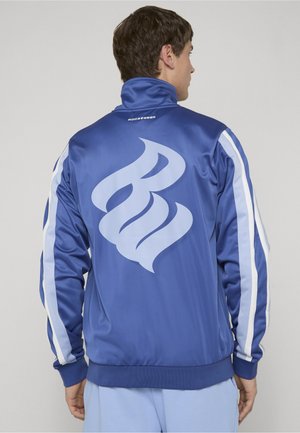 Homme portant un blouson de survêtement bleu avec des bandes blanches sur les manches et un grand logo abstrait dans le dos, tourné dos à un fond uni.
