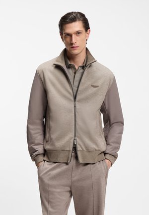 Junger Mann mit zurückgegtem Haar, trägt eine beige Jacke mit Reißverschluss über einem passenden Poloshirt und Hose, Hände in den Taschen, neutraler Gesichtsausdruck.