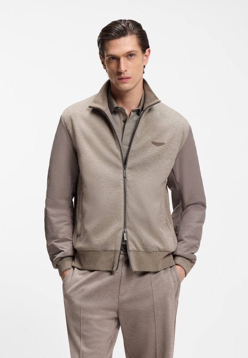 Jeune homme aux cheveux plaqués en arrière portant une veste beige zippée sur un polo et un pantalon assortis, mains dans les poches, expression neutre.