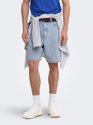 Mężczyzna ubrany w jasnoniebieskie dżinsowe szorty, białe trampki, białe skarpetki, niebieski t-shirt i pasiastą koszulę związaną wokół talii.