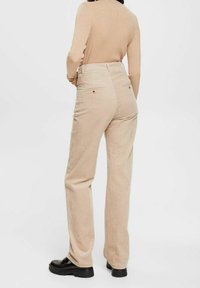 Person wearing beige ribbed pants, beige long-sleeve top, and black platform shoes standing against a plain background.