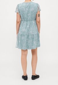 Femme en robe courte fleurie bleu clair avec manches volantées et chaussures Mary Jane noires, debout de dos, montrant un tatouage sur son bras gauche.