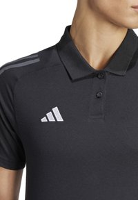 Polo nero con colletto a bottoni, con strisce grigie sulle spalle e logo Adidas bianco. Realizzato in tessuto testurizzato.