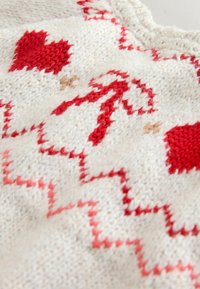 Knit textile with a cream base featuring red hearts and zigzag patterns. Soft texture with intricate details in contrasting yarn colors.