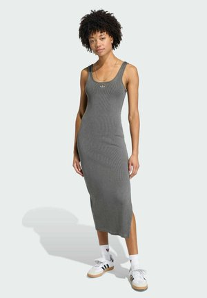 Donna in piedi che indossa un abito midi grigio aderente senza maniche con spacco laterale, calzini bianchi Adidas e sneakers bianche Adidas con strisce nere.