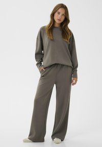 Woman wearing loose grey sweatshirt and wide-leg pants with white sneakers, standing with one hand in pocket on plain white background.