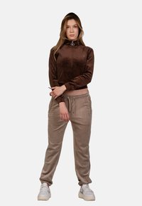 Giacca zip-up in velour marrone con colletto alto e polsini a costine abbinata a joggers beige chiaro con vita regolabile e polsini elasticizzati. Sneakers bianche.