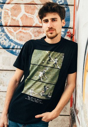 Zwarte katoenen t-shirt met drie grafische panelen van voetbal scènes tegen een groene grasachtergrond; bevat witte tekst onderaan.