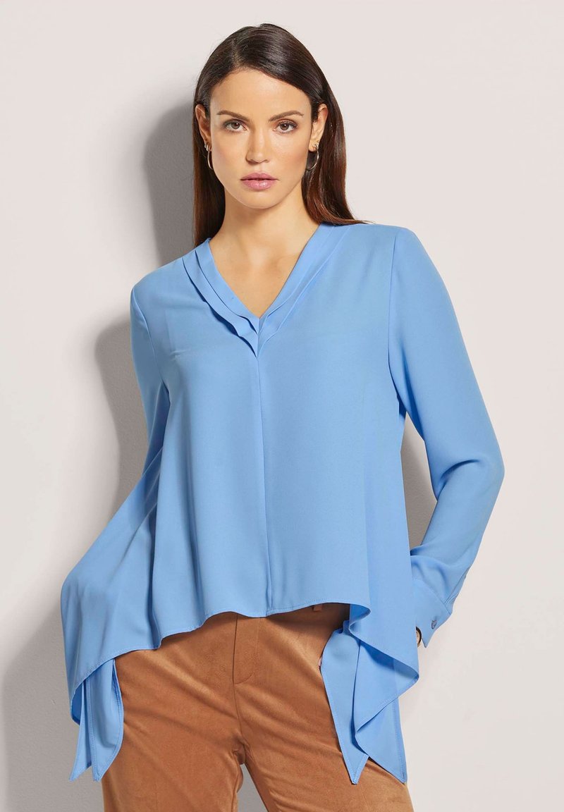 Helle blaue Bluse mit V-Ausschnitt, langen Ärmeln und asymmetrischem Saum. Hergestellt aus glattem Stoff mit fließender Textur.