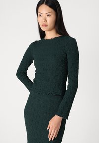 Green textured long-sleeve top with a scalloped neckline, paired with a matching textured pencil skirt. The fabric has a crinkled appearance.