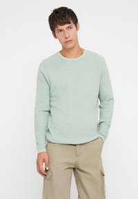 Ανοιχτό πράσινο, υφαντό πουλόβερ με στρογγυλή λαιμόκοψη και μακριά μανίκια, σε συνδυασμό με μπεζ παντελόνια cargo. Απλός σχεδιασμός χωρίς ορατά μοτίβα.