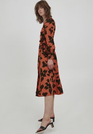 Langärmliges Kleid in Rost mit einem schwarzen Blumenmuster, tailliertem Bund und knielangem Rock. Kombiniert mit schwarzen, hohen Sandalen.