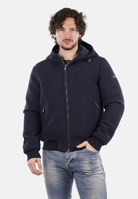 Giacca con cappuccio blu navy, con chiusura a zip sul fronte, polsini a coste e tasche laterali. Realizzata in tessuto testurizzato con una fodera interna liscia.