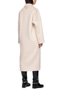Long, beige wool coat with a collared neck, dropped shoulders, and back seam detail. Worn over black, glossy knee-high boots with gold accents.