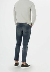 Dżinsy w ciemnym odcieniu o slim fit, z tylnymi kieszeniami i subtelnym przetarciem. Noszone z jasnoszarym swetrem i czarnymi sneakersami.