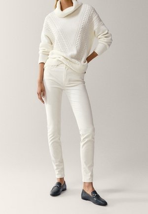 Modèle portant un pull à col roulé en maille torsadée blanche, un pantalon slim blanc et des mocassins noirs, debout sur un fond neutre.