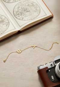 Guldarmband med glob- och flygplansberlocker, delikat kedja, placerad på en beige tygyta, bredvid en läderinbunden bok och en kamera.