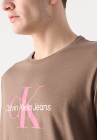 Pruun puuvillane t-särk, millel on suur roosa "CK" ja tekst "Calvin Klein Jeans". Ümmargune kaelus, sile tekstuur ja lühikesed varrukad.