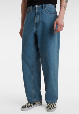 Persona che indossa jeans blu larghi e scarpe slip-on a quadretti bianche e nere, in piedi davanti a uno sfondo semplice.