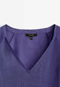 Haut à col en V, manches courtes, de couleur pourpre profonde. Fabriqué en tissu léger avec une finition légèrement texturée. Étiquette visible à l'intérieur du col.