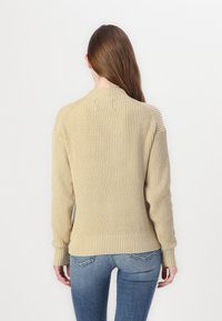 Beżowy sweter z prążkowanego dzianiny z długimi rękawami i okrągłym dekoltem, charakteryzujący się teksturowanym wykończeniem i dopasowanymi mankietami, w połączeniu z niebieskimi dżinsami.