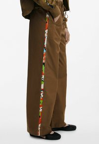 Hnedé nohavice s širokými nohavicami majú farebný vzorovaný bočný pruh. Vyrobené z hladkej tkaniny s voľným strihom, obsahujú bočné vrecká.