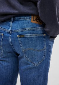 Jeans de denim albastru cu o textură netedă, având o etichetă din piele maro pe talie și o etichetă mică neagră pe buzunarul din spate.