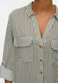 Femme portant une chemise à rayures verticales vert olive et blanches, avec des poches poitrine à rabat et des manches retroussées.