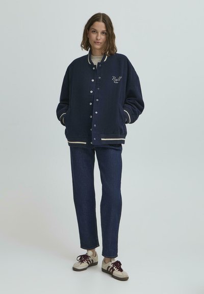 Jachetă de tip varsity navy cu manșete și tiv alb cu nervuri, având un logo mic. Purtată peste un pulover ușor și asortată cu blugi închisi la culoare și adidași.