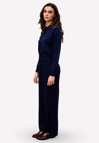 Pull en maille bleu marine avec un col zippé moitié, associé à un pantalon large. Tissu texturé et coupe décontractée, complété par des mocassins marron.