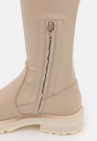 Beige læder knæhøj støvle med en sidelukning og kontrasterende hvid syning. Har en kraftig cremefarvet sål for godt fodfæste.